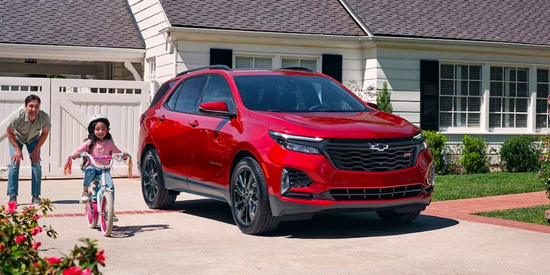 A father helping his daughter ride a bicycle in their driveway where their new cherry red 2024 Chevrolet Equinox sits.