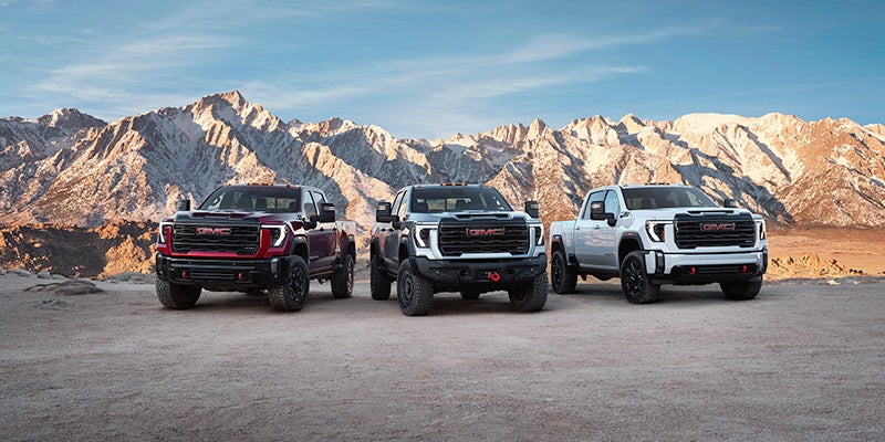 The image features three GMC trucks parked in front of a mountain range. The trucks are positioned side by side, with one being red, one being black, and the third one being white. The mountain range serves as a stunning backdrop for the vehicles, emphasizing their rugged and powerful nature. The trucks are likely showcasing their capabilities and features, making them an attractive choice for those who need a reliable and versatile vehicle for their outdoor adventures.
